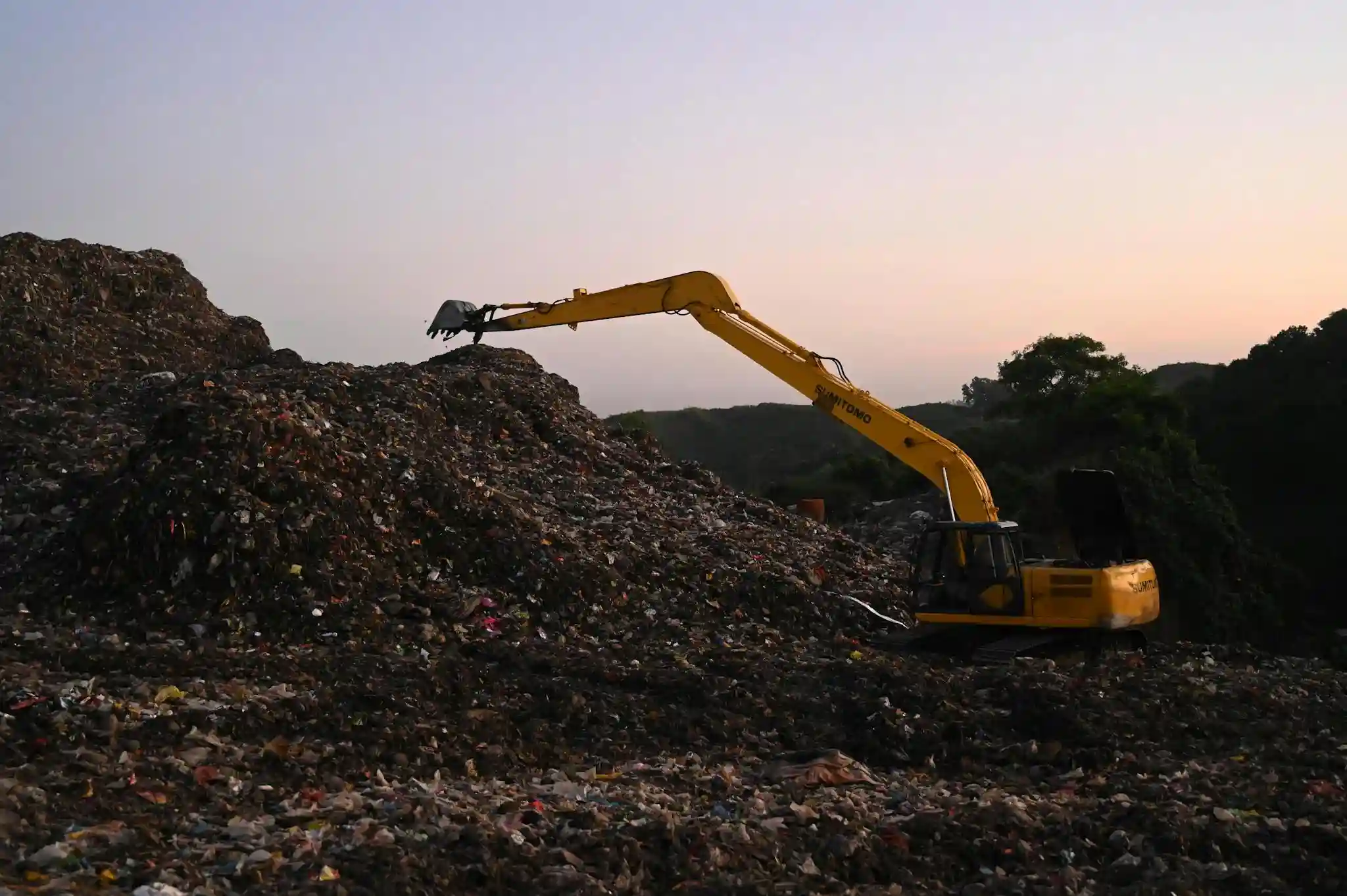 Yellow excavator With Adblue / DEF Delete Emulator Fitted operates amidst piles of waste in a Bangladesh landfill at sunset.